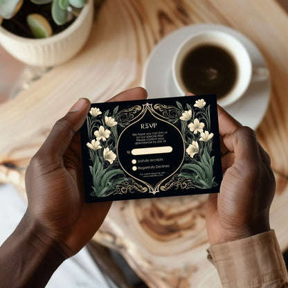 Person holding a decorative dark art nouveau wedding rsvp card with floral design on a wooden table with a cup of coffee.